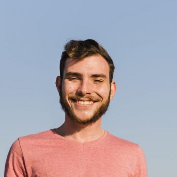 portrait-of-a-smiling-young-man-standing-on-the-beach-against-blue-sky